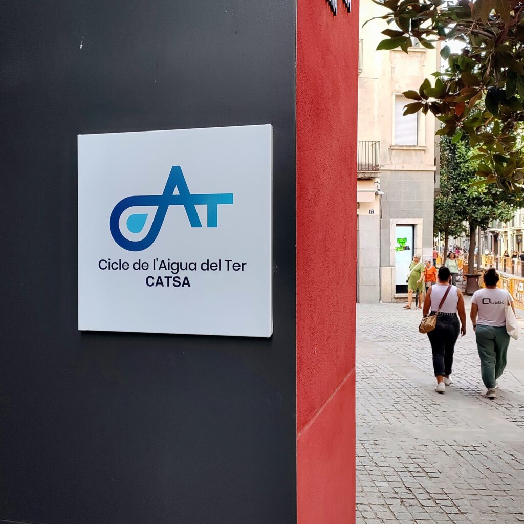 Placa amb el logotip de Cicle de l'Aigua del Ter (CATSA) a la façana d'un edifici, amb un carrer de vianants al costat
