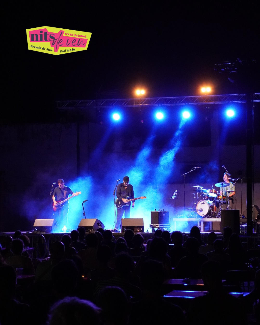 Un trio de baix, guitarra i bateria toca de nit entre fum i llums blaves, amb el públic en silueta, al festival Nits de Veu