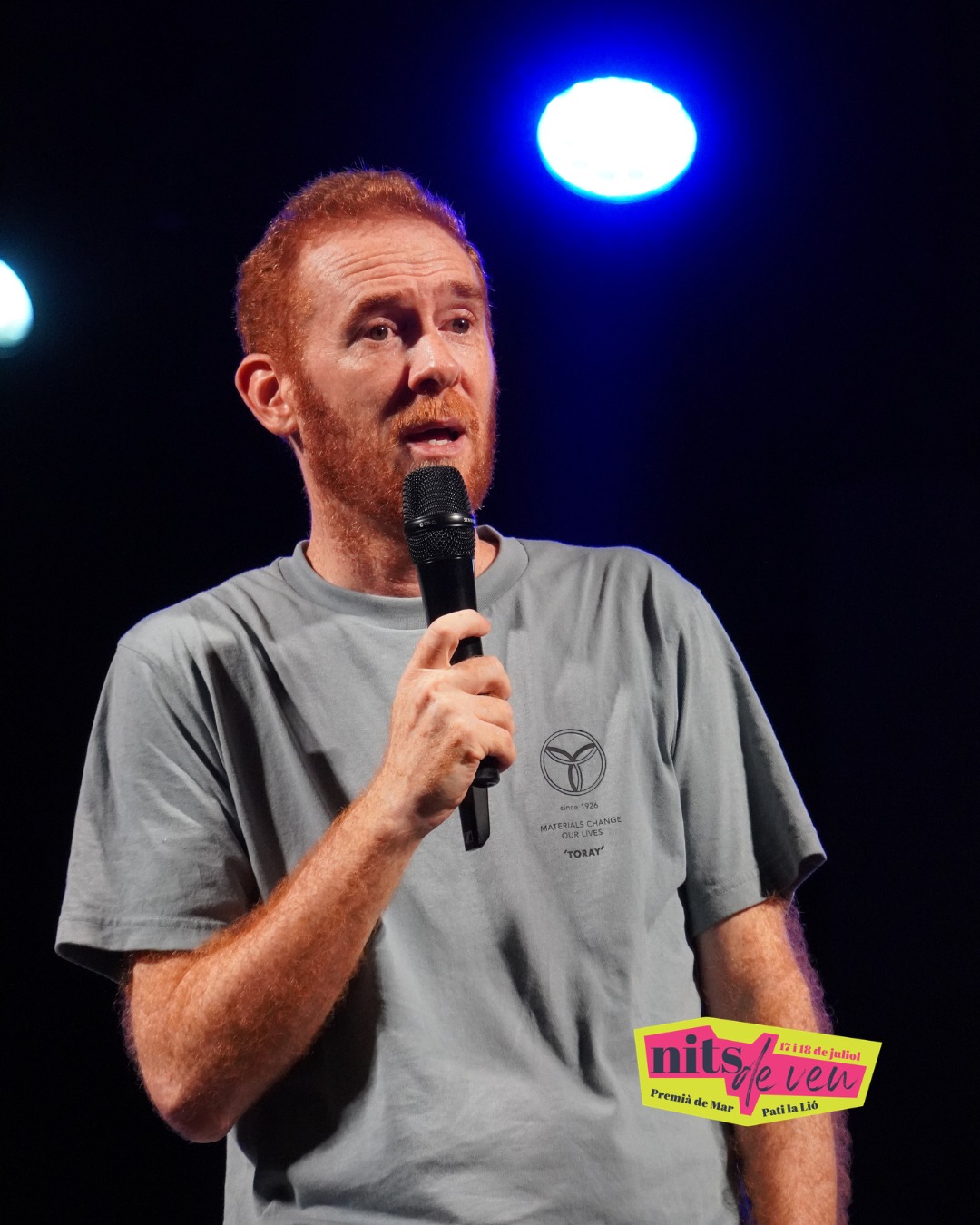 Un humorista pèl-roig parla al micròfon sota un focus blau a l'escenari del festival Nits de Veu